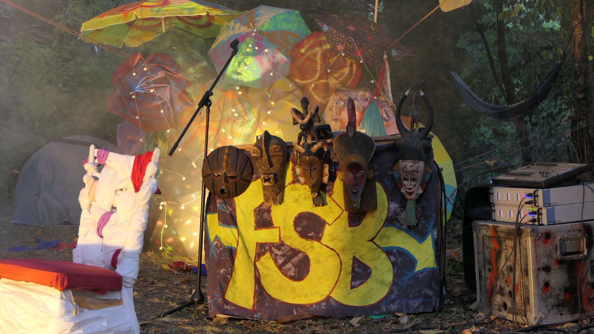 Bout de décor du spectacle, il y a une table décorée d'un tissu avec un tag FSB dessus, ainsi que des masques. En arrière plan, une structure composée de parapluies. Jeu de lumière entre les deux.