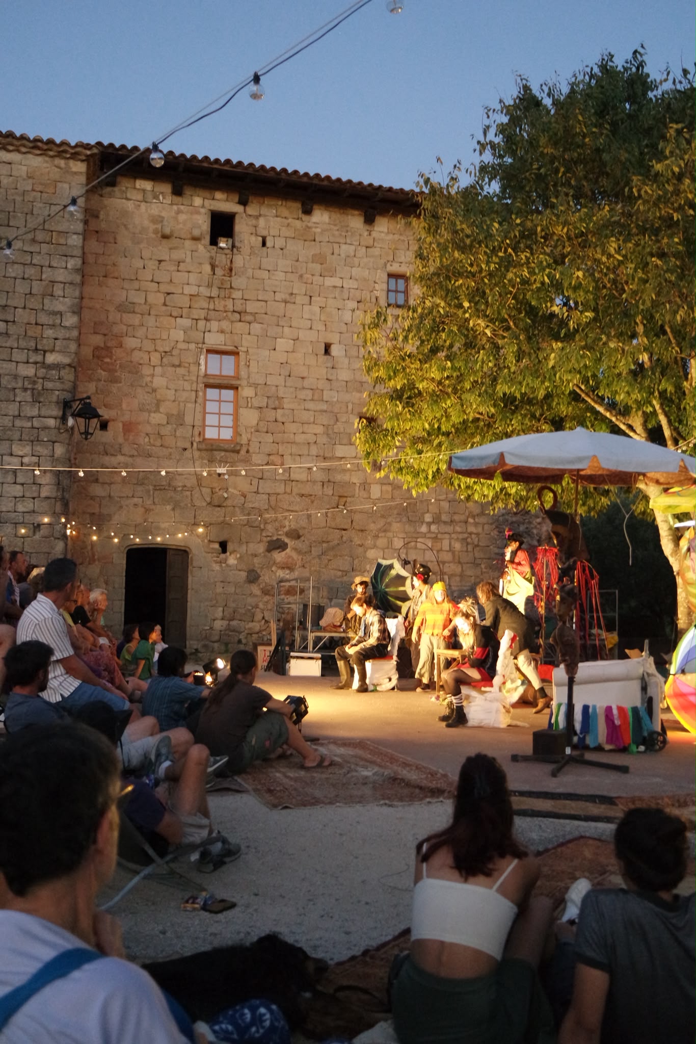 Spectacle joué dehors à la tombée du jour, on voit les comédiens dans la lumière, le public à gauche et un château en arrière plan.